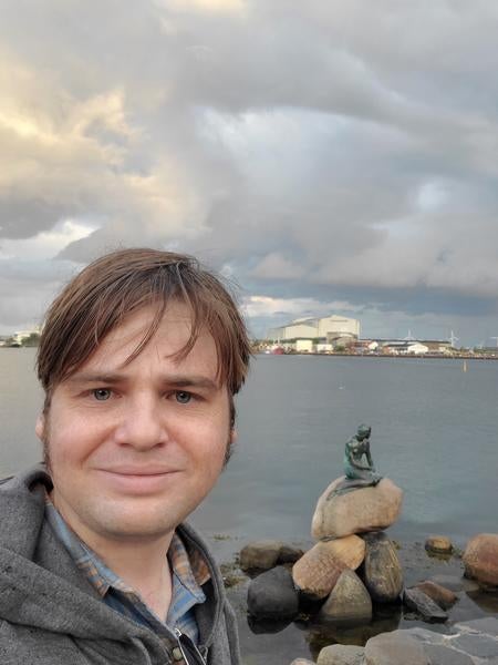 Bryan from the shoulders up, standing in front of the Hans Christian Anderson mermaid statue in Copenhagen harbor. 