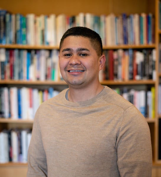 Photo of Carlos from the elbows up, smiling, in front of a bookcase