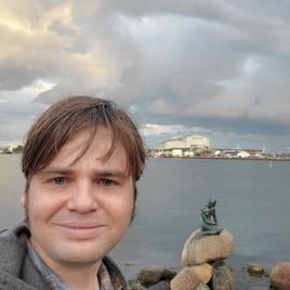 Bryan from the shoulders up, standing in front of the Hans Christian Anderson mermaid statue in Copenhagen harbor. 
